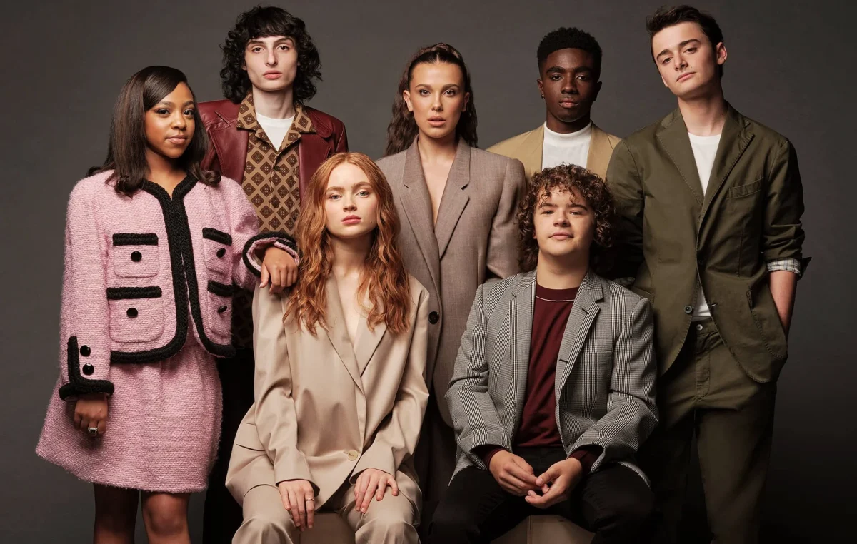 A group portrait of the core Stranger Things cast posing together in formal, fashion-forward outfits against a neutral studio backdrop.
