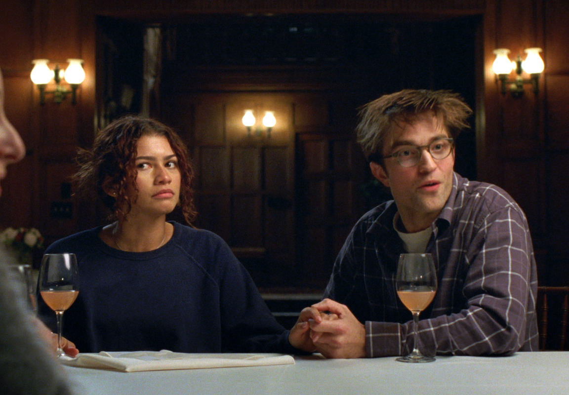Actors Zendaya and Robert Pattinson sitting together at a table with glasses of wine in a scene from the film "The Drama."